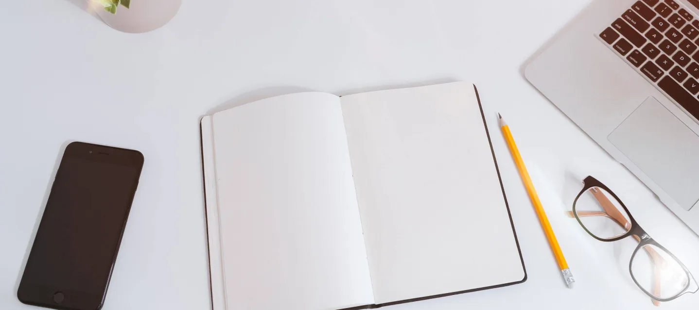 Open notebook, pencil, phone, and laptop on a white desk.